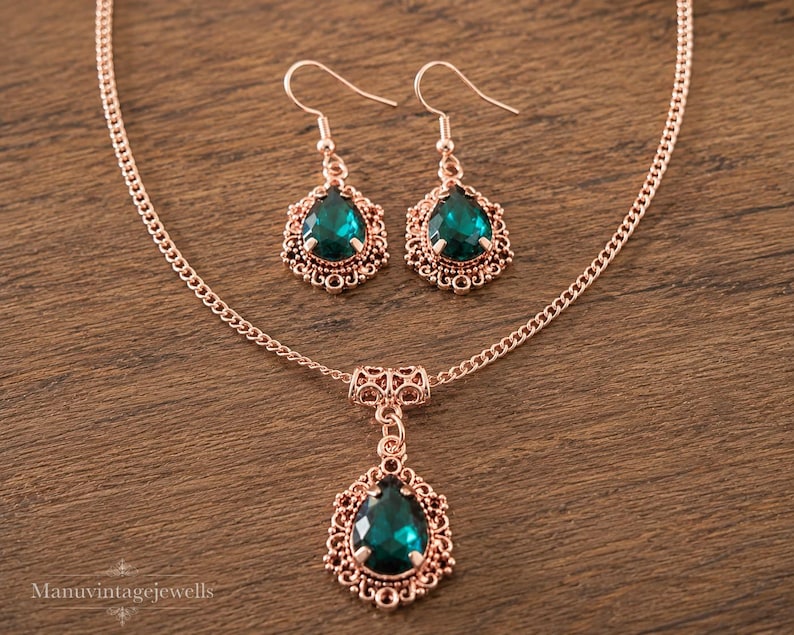 Puede incluir: Un conjunto de collar y pendientes en oro rosa con cristales verde esmeralda en forma de l&aacute;grima. El colgante y los pendientes est&aacute;n enmarcados por un intrincado trabajo de filigrana met&aacute;lica. El collar incluye una cadena fina y ganchos.