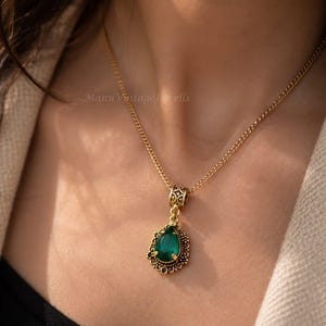 Puede incluir: Un collar de tono dorado con un colgante de piedra preciosa verde en forma de l&aacute;grima. El colgante est&aacute; engastado en un marco de metal dorado con filigrana ornamentada y una anilla decorativa a juego, suspendido de una fina cadena.