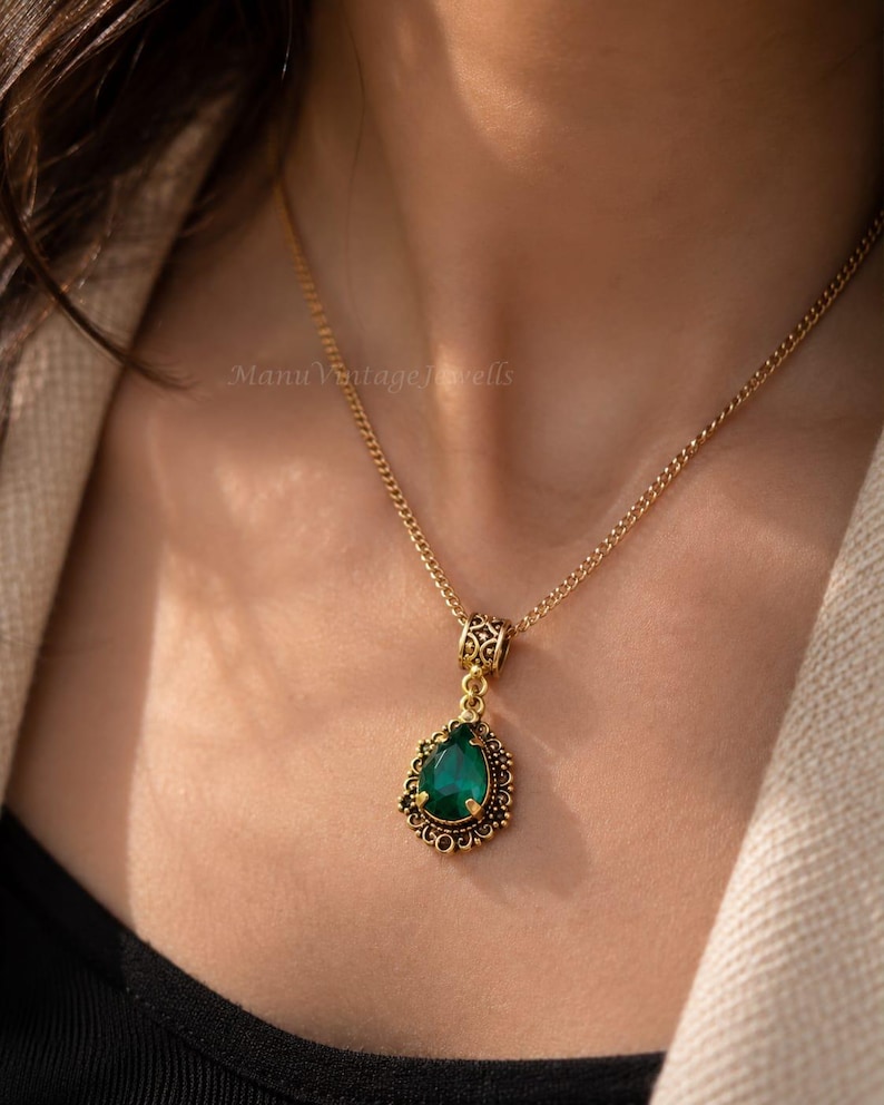 Puede incluir: Un collar de tono dorado con un colgante de piedra preciosa verde en forma de l&aacute;grima. El colgante est&aacute; engastado en un marco de metal dorado con filigrana ornamentada y una anilla decorativa a juego, suspendido de una fina cadena.