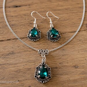 Puede incluir: Un conjunto de joyer&iacute;a en tono plateado que consta de un collar y un par de pendientes colgantes. Cada pieza presenta una piedra preciosa verde oscuro facetada en forma de pera, engastada en un marco plateado ornamentado y filigranado.
