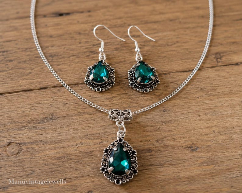 Puede incluir: Un conjunto de joyer&iacute;a en tono plateado que consta de un collar y un par de pendientes colgantes. Cada pieza presenta una piedra preciosa verde oscuro facetada en forma de pera, engastada en un marco plateado ornamentado y filigranado.