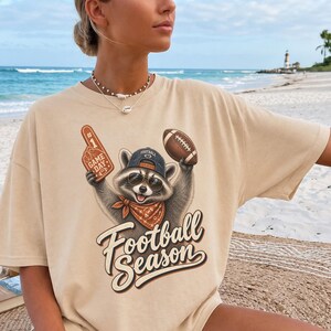 Peut inclure: Un t-shirt beige à manches courtes orné d'un motif représentant un raton laveur portant des lunettes de soleil, une casquette et un bandana orange. Le raton laveur tient un doigt en mousse et un ballon de football. Le texte Football Season est imprimé en dessous.