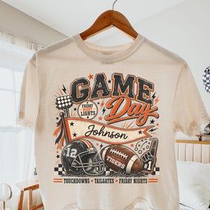 May include: A cream-colored short-sleeve t-shirt featuring a graphic print with the text "GAME Day", "FRIDAY NIGHT LIGHTS", "Johnson", and "TOUCHDOWNS TAILGATES FRIDAY NIGHTS". The design includes a football helmet, a football, and a foam finger.