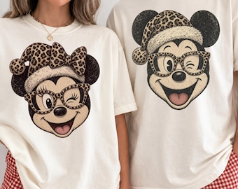 May include: Two off-white crew neck t-shirts featuring graphic prints of cartoon mouse faces. One shirt displays a mouse with a leopard print bow and hat, the other a mouse with a leopard print hat. Both wear leopard print glasses.