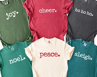 May include: A collection of six short-sleeved crew neck t-shirts in various colors including olive green, red, charcoal gray, teal, and cream. Each shirt features a single word or phrase printed in a simple font followed by a period.