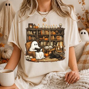Könnte beinhalten: Ein cremefarbenes T-Shirt mit einem Grafikdruck eines Geistes, der in einem Sessel sitzt und ein Buch vor einem Bücherregal liest, das mit Büchern, Kerzen, Kürbissen und einer schwarzen Katze gefüllt ist. Das Design enthält Totenköpfe.