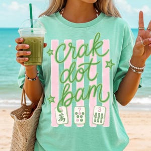 May include: A mint green short-sleeve t-shirt featuring a graphic print of vertical pink stripes, green stars, and the cursive text "crak dot bam". Below the text are three mahjong tiles displaying the numbers 5, 7, and 3 with corresponding symbols.