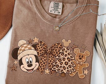 May include: A brown short-sleeve t-shirt featuring a graphic of Mickey Mouse wearing a checkered beanie, a large leopard print bow, a leopard print Christmas tree, and a gingerbread man. The shirt has a Comfort Colors brand label.