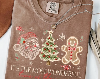May include: A brown short-sleeve t-shirt featuring a graphic of Santa Claus, a Christmas tree, and a gingerbread man. The text reads, "IT'S THE MOST WONDERFUL time of the year." A silver chain necklace rests on the neckline of the shirt.