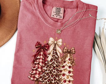 May include: A dusty rose short-sleeve t-shirt with a Comfort Colors label at the neck. The graphic features three stylized Christmas trees topped with large bows, showcasing red plaid, leopard print, and polka dot patterns with gold stars.