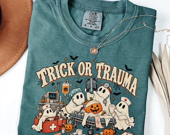 May include: A teal short-sleeved t-shirt featuring a graphic of ghosts dressed as medical staff with pumpkins and the text "TRICK OR TRAUMA". The shirt has a crew neckline and a Comfort Colors brand label printed on the inside of the collar.