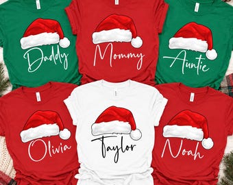 May include: A collection of six short-sleeved crew neck t-shirts in red, green, and white. Each shirt features a graphic of a red Santa hat with white trim and the printed names or titles Daddy, Mommy, Auntie, Olivia, Taylor, and Noah.