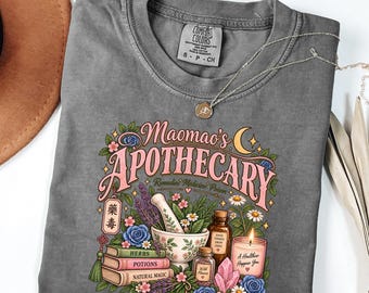 May include: A charcoal gray t-shirt featuring a graphic print with the text Maomao's APOTHECARY, Remedies Medicine Poison, Herbs, Potions, Natural Magic, Good Things Grow Here, and A Healthier Happier You, alongside illustrations of a mortar and pestle, books, and a candle.