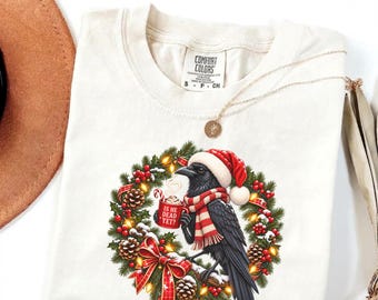 May include: A cream-colored t-shirt featuring a graphic of a black crow wearing a red and white Santa hat and scarf. The crow holds a red mug with the text IS HE DEAD YET? inside a Christmas wreath with pinecones, red berries, and lights.