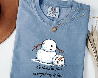 May include: A blue crew neck t-shirt featuring a graphic of a snowman with a detached head resting on the ground. The text below the snowman reads, "it's fine, i'm fine everything is fine." The shirt has a Comfort Colors brand label.