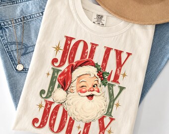 May include: An off-white t-shirt featuring a graphic of a winking Santa Claus wearing a red hat with holly berries. The text "JOLLY" appears in red above and below the Santa illustration, accented by gold starbursts and green lettering.
