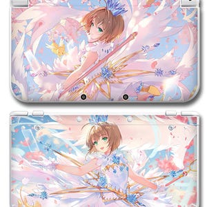 May include: A white handheld gaming console featuring a full-color anime illustration of a character with short brown hair, green eyes, and large white wings. The character wears a white dress with blue accents and holds a staff amidst pink petals.