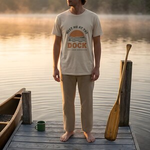 May include: A beige short-sleeve t-shirt featuring a graphic print of a sunset over water with the text "MEET ME AT THE DOCK" and "EST. LAKE WEEKENDS" in dark brown lettering. The shirt has a crew neckline and a standard fit, made of cotton fabric.