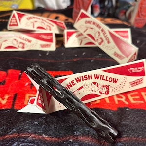 Peut inclure: Un objet noir texturé ressemblant à une branche posé sur une boîte triangulaire blanche et rouge marquée ONE WISH WILLOW. L'emballage présente des illustrations de deux visages, le texte AMAZE YOUR FRIENDS et la phrase You only get ONE WISH.