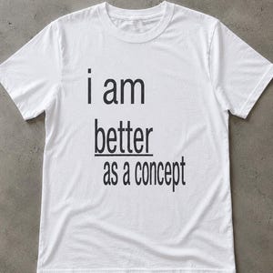 May include: A white short-sleeved t-shirt featuring black text printed on the front. The text reads i am better as a concept, with the word better underlined. The garment has a crew neckline and a standard fit, constructed from plain white fabric.
