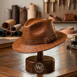 Puede incluir: Un sombrero fedora marrón con un patrón texturizado de piel de cocodrilo y una banda de cuero a juego con una hebilla de metal plateado. El sombrero se exhibe sobre un soporte de madera oscura con un logotipo circular y el texto WL Wear Legacy.