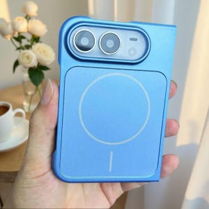 Puede incluir: Una funda para smartphone de color azul claro que presenta un anillo de carga magnético circular prominente en el panel posterior. La funda incluye un módulo de cámara rectangular elevado con dos lentes circulares y un pequeño flash.