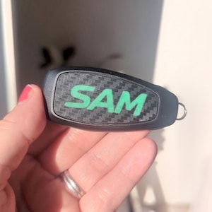 May include: A black key fob cover featuring a carbon fibre texture and the name SAM printed in bright green capital letters across the centre. The accessory includes a small metal ring attachment on one side for securing to a keychain.