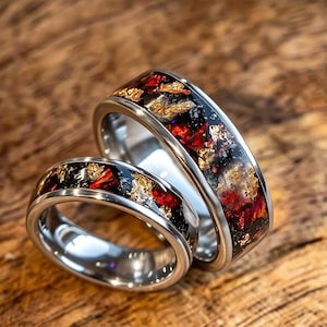 May include: Two silver-toned metal rings featuring a central inlay of crushed red, black, and gold-colored flakes. The rings have a polished, reflective finish on the inner bands and a smooth, rounded exterior profile showcasing the colorful inlay.