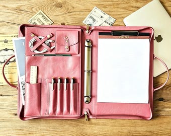 May include: A pink leatherette zippered portfolio organizer featuring a three-ring binder, a notepad labeled Veritas, multiple pen slots, a small snap-closure pocket, a cable organizer, and a vertical sleeve for documents, all arranged in a structured interior.