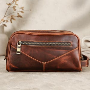 May include: A rectangular brown leather toiletry bag featuring a horizontal zippered front pocket with a metallic pull tab. The bag has visible stitching along the seams, a structured shape, and a small leather side handle for carrying purposes.