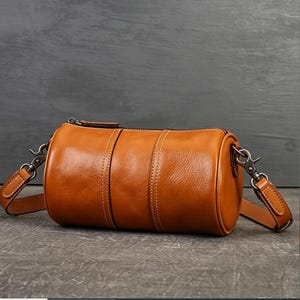 May include: A cylindrical tan leather barrel bag featuring a top zip closure, white contrast stitching, and dark metal hardware. The bag includes a detachable shoulder strap with swivel lobster clasps attached to each side of the main body.