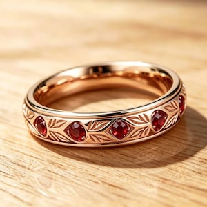 May include: A rose gold band ring featuring a repeating pattern of engraved leaves and small, round, faceted red gemstones set at regular intervals around the entire circumference of the metal band, creating a continuous decorative design.