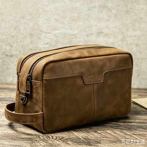 May include: A rectangular brown leather wash bag featuring a side handle, two top zips with dark metal pulls, and the letters MNT embossed on the front panel. The bag has visible stitching along the edges and a smooth, matte finish.