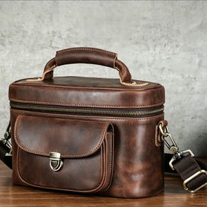 May include: A dark brown leather bag with a top handle and a front pocket featuring a metal clasp closure. The bag includes a zippered main compartment and detachable brown fabric shoulder straps with metal hardware clips on both sides.