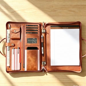 May include: A brown leather-like portfolio organizer featuring a zippered closure, multiple card slots, a small snap-button pocket, pen loops, a smartphone sleeve, and a metal ring binder holding a lined notepad with the word Veritas printed at the top.