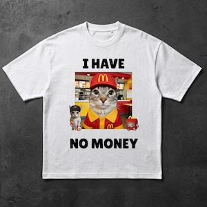 Op de afbeelding: Een wit T-shirt met korte mouwen en een grafische print van een kat in een rood-geel uniform en pet in een restaurant. De tekst I HAVE NO MONEY staat in het zwart gedrukt boven en onder de afbeelding van de kat en twee kleinere katten.