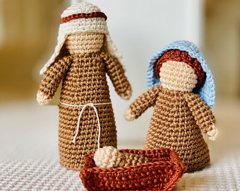 May include: Crocheted nativity scene featuring three figures and a manger. The figures include a man in a brown robe and white head covering, a woman in a blue head covering, and a baby in a brown manger. The figures are made of tan and brown yarn.