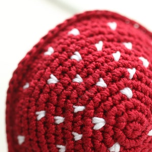 May include: Close-up of a red crocheted hat with white heart-shaped details.