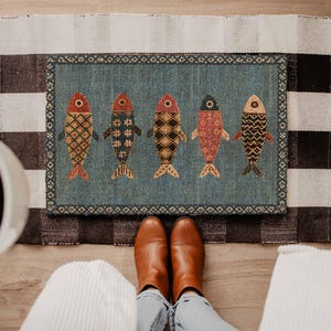 May include: A rectangular rug featuring a blue background with five stylized fish arranged in a row. Each fish displays a unique geometric pattern in shades of red, orange, black, and cream. The rug is bordered by a decorative dark patterned trim.