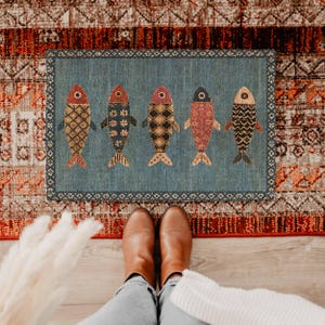 May include: A rectangular blue doormat featuring a decorative border and five stylized fish arranged in a horizontal row. Each fish displays a unique pattern in shades of red, orange, black, and cream, set against a textured blue background.