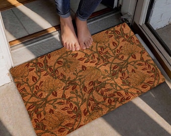 May include: A rectangular doormat featuring a repeating pattern of large, stylized orange flowers and dark green leaves on a textured, light brown coir background. The mat is positioned on a floor surface with a visible door frame threshold.