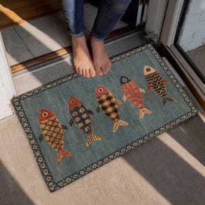 May include: A rectangular doormat with a blue background and a decorative border featuring five fish arranged in a row. Each fish displays a unique pattern, including diamonds, stars, and solid colors in shades of orange, red, and black.