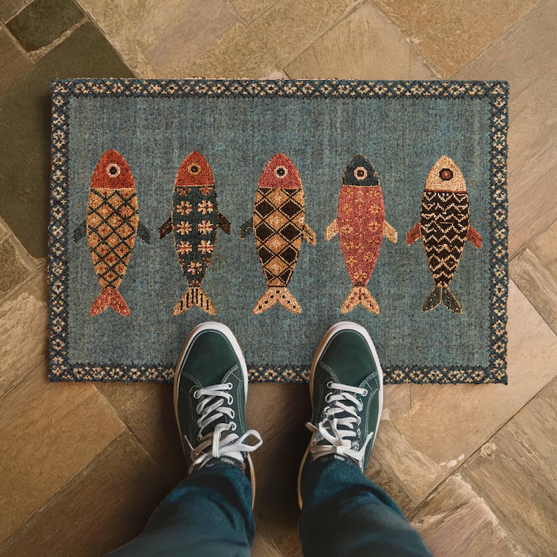 May include: A rectangular doormat with a blue background featuring five stylized fish arranged in a row. Each fish displays a unique geometric pattern in shades of red, orange, black, and cream. The mat has a dark blue border with a repeating pattern.
