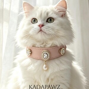 Puede incluir: Un collar para mascotas de color rosa claro con tres adornos circulares incrustados de cristales y un dije central de perla en forma de lágrima. El texto KADAPAWZ - Interchangeable Charms Pet Collar - está impreso en negro en la parte inferior.