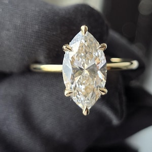 Könnte beinhalten: Ein Ring mit einem Diamanten im Marquise-Schliff, gefasst in eine dünne Ringschiene aus Gelbgold. Der Mittelstein wird von sechs Krappen gehalten und hat eine spitz zulaufende ovale Form mit zahlreichen Facetten, die das Licht reflektieren.