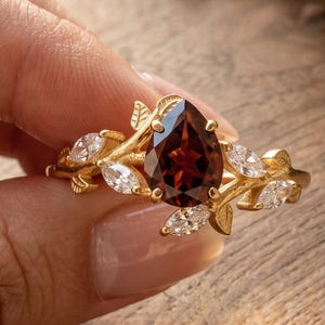 May include: A gold ring featuring a central pear-shaped dark red garnet gemstone. The band is designed with a vine-like motif, incorporating small gold leaves and several marquise-cut clear white stones arranged along the sides of the setting.