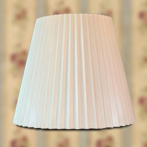 May include: A white, conical lampshade featuring a uniform, vertical pleated texture throughout its surface. The lampshade has a wide base that tapers toward a narrower top, creating a clean, structured silhouette for a lighting fixture.