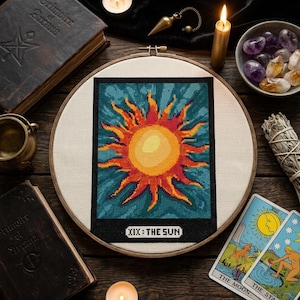 May include: A circular wooden embroidery hoop containing a cross-stitch design of a bright yellow and orange sun with radiating flames against a dark teal background, featuring the text XIX: THE SUN in a black rectangular border at the bottom.