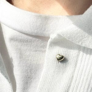 May include: A small, gold-colored heart-shaped pin is affixed to a white knit top. The pin is centered on the fabric, adding a subtle touch of elegance. The top has a crew neck and is made of a soft, textured material.