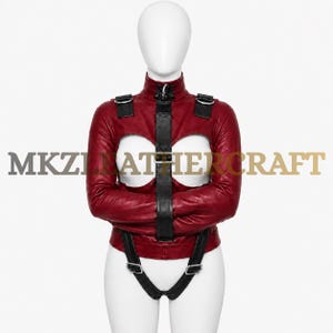 May include: A deep red leather jacket featuring long sleeves, a high collar, and two large circular chest cutouts. The garment includes black leather straps with silver-toned metal buckles and D-rings, and the text MKZ LEATHERCRAFT is visible.